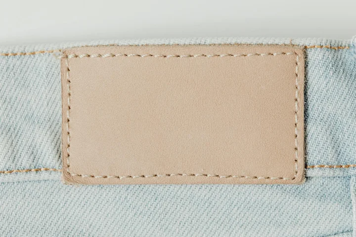 Close-up of light-wash denim fabric with a beige leather patch stitched onto the waistband.
