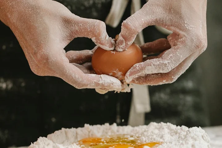 Hands dusted with flour crack an egg over a bowl of flour in a kitchen.