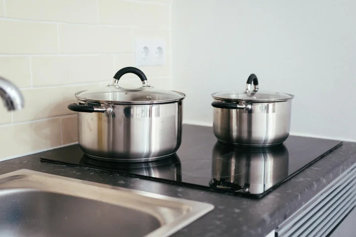 Two stainless steel pots with lids on a black glass electric stove top in a modern kitchen, with a sink and backsplash visible in the background.