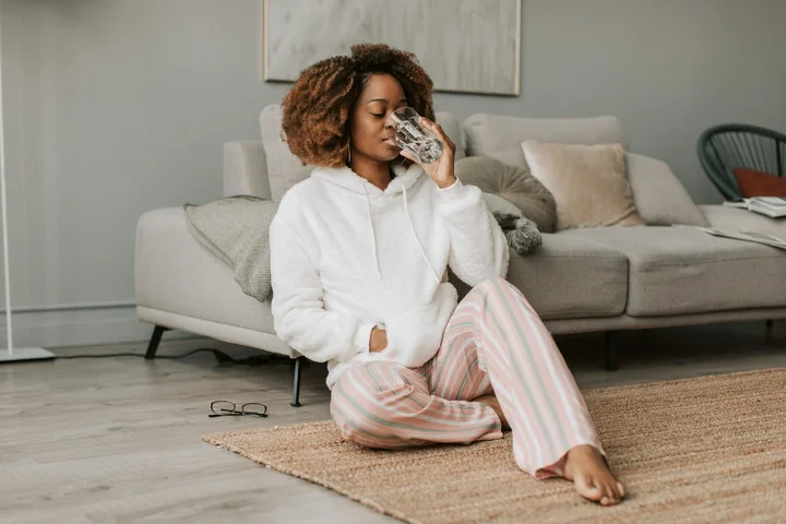 A woman sits on a beige rug in a living room, wearing a white hoodie and pink striped pants, holding a tissue to her nose. Glasses lie on the rug beside her, with a sofa and cushions in the background.