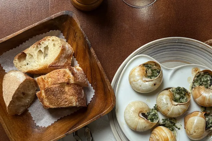 Rustic wooden table featuring a basket of sliced bread and a plate with small round baked onions topped with herbs.