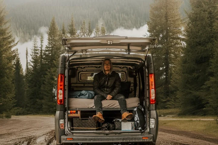 Person sitting at the open rear of a cargo van on a dirt road in a forested area, gear visible inside the van.
