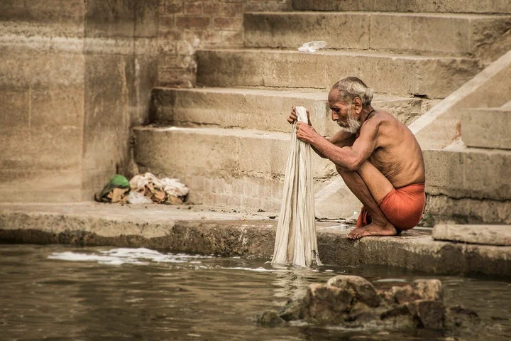 An elderly man, shirtless and wearing red shorts, sits by stone steps and rinses a cloth in a body of water.
