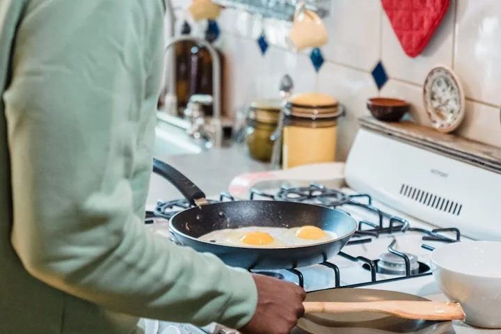 Person wearing a green sweater cooking eggs in a frying pan on a gas stove in a bright kitchen with a tiled backsplash and kitchen tools in the background.
