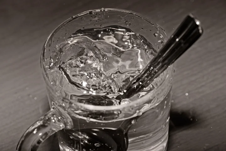 Close-up of a glass mug filled with water and a spoon inside on a dark surface, illustrating an immediate spill.