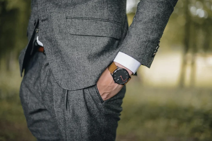 Close-up of a person wearing a gray suit with a white shirt cuff and a wristwatch, standing outdoors with a blurred park background.