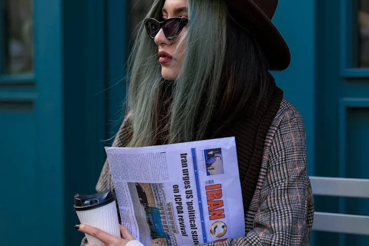 Person with green-tinted hair wearing sunglasses and a hat, reading a newspaper outdoors