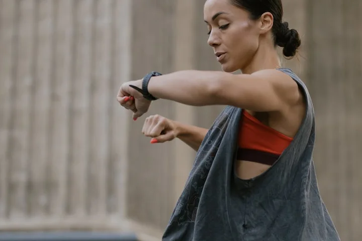 Athlete wearing a gray sleeveless top and a red sports bra checks her smartwatch outdoors near a railing.