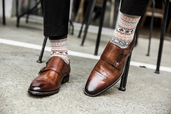 Close-up of brown leather buckle monk-strap shoes on a sidewalk, paired with patterned knit socks.