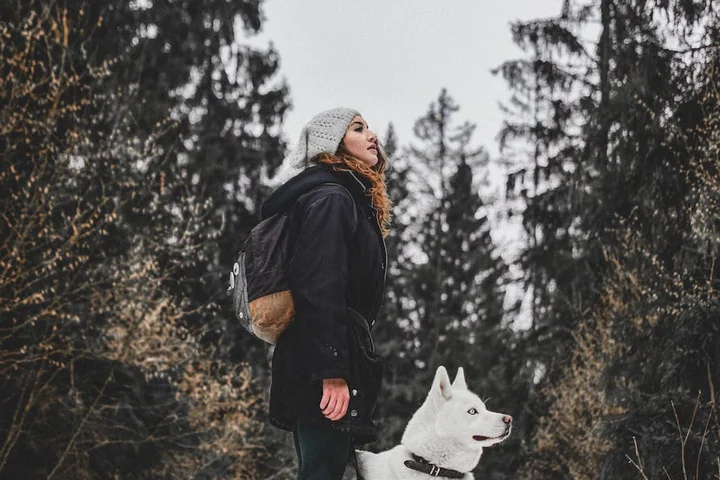 Person wearing a dark coat and gray knit beanie with a backpack standing in a snowy forest beside a white dog