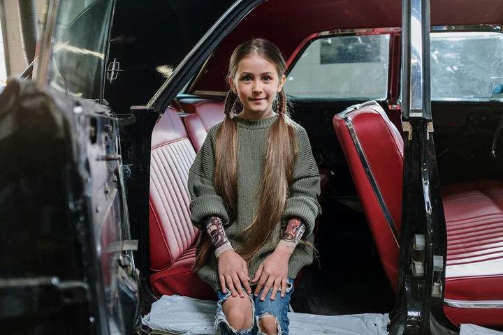 Young girl with long braids sits inside an open vintage car with red leather seats.