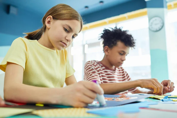 Two children sit at a table filled with colorful craft supplies, using glue sticks during a craft project.
