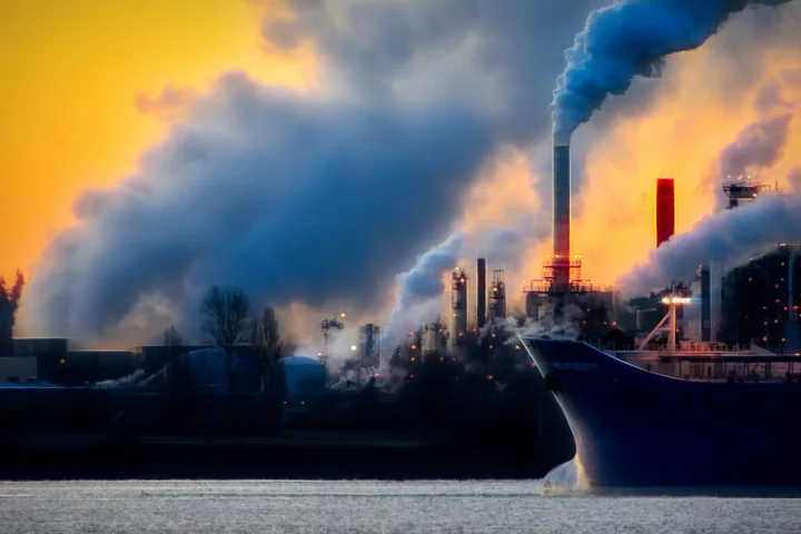 Industrial harbor scene with large smokestacks emitting thick smoke over a body of water at sunset.