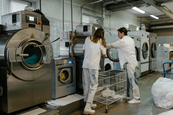 Two technicians in white lab coats inspect commercial washing machines in an industrial laundry facility, preparing to tackle stubborn odors.