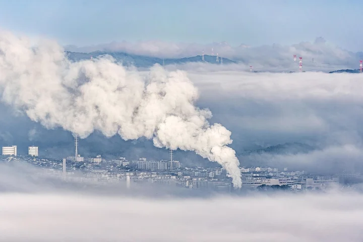 Industrial smokestacks emitting white plumes over a foggy, industrial landscape.