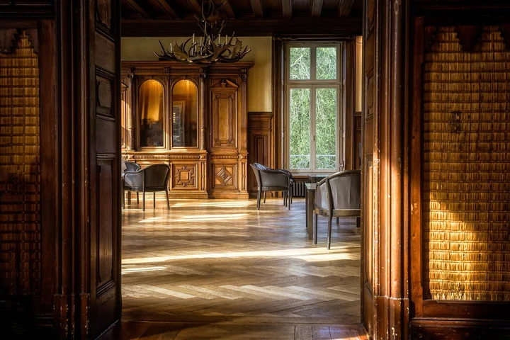 A sunlit classical room with warm wooden parquet floors, ornate wood paneling, and vintage furniture
