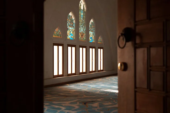 Open doorway to a sunlit interior with stained-glass windows and a blue patterned carpet.