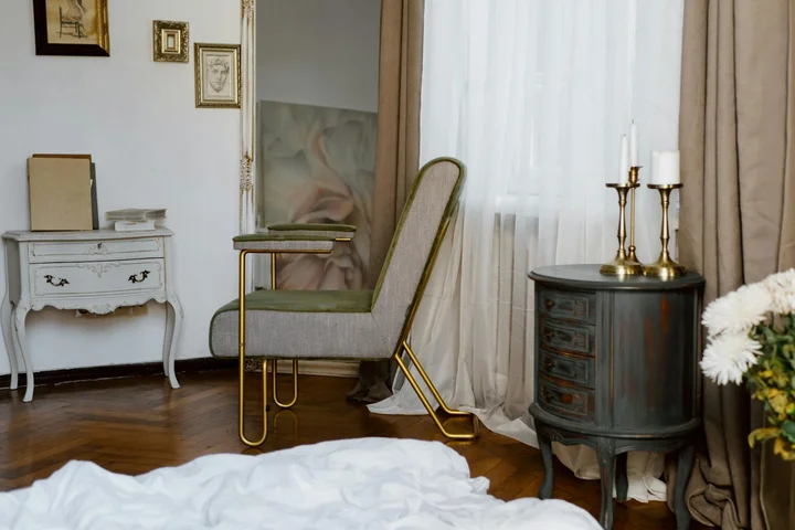 A stylish living space with a white dresser, a green upholstered chair with a gold metal frame, a dark wood side table with candlesticks, and sheer curtains beside a bed with white linens.