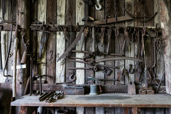 A rustic workshop wall with an assortment of old hand tools hanging from a pegboard above a weathered wooden workbench.