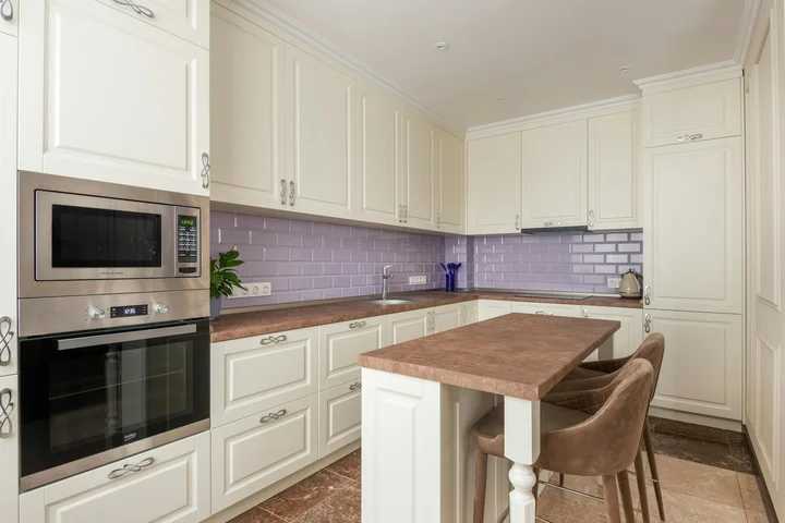 Bright white kitchen with a built-in stainless steel microwave, white cabinets, lavender subway tile backsplash, and a wooden island with two brown chairs.