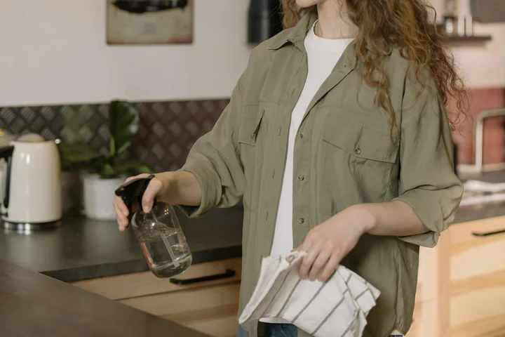 Person wearing an olive-green shirt cleaning a kitchen countertop with a spray bottle and microfiber cloth.
