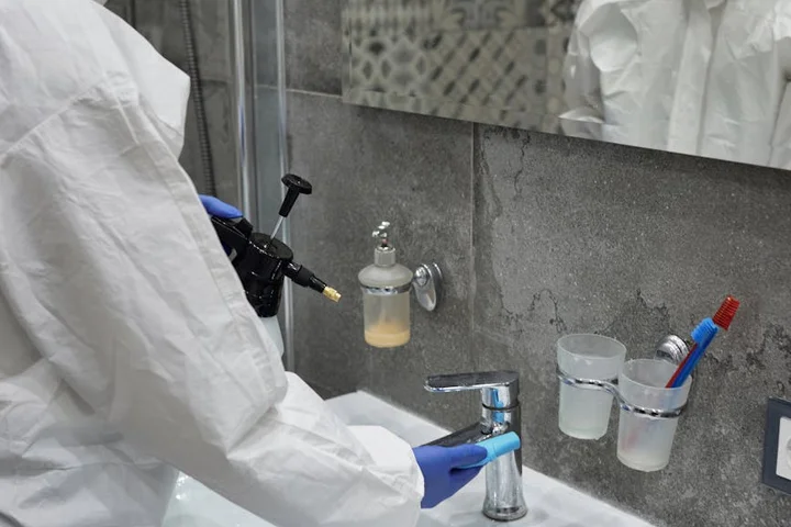 A lab technician wearing a white protective suit and blue gloves uses a spray bottle to clean a laboratory sink, faucet, and glassware mounted on a gray tiled wall.