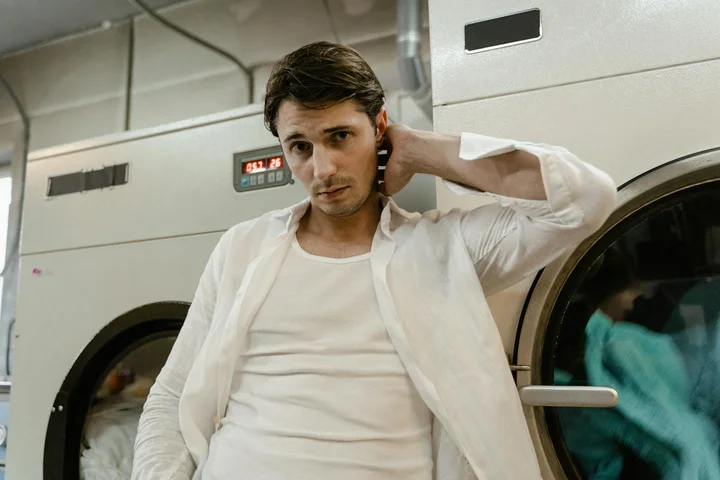 A man in a white shirt stands beside a front-loading washing machine in a laundromat, looking at the camera.