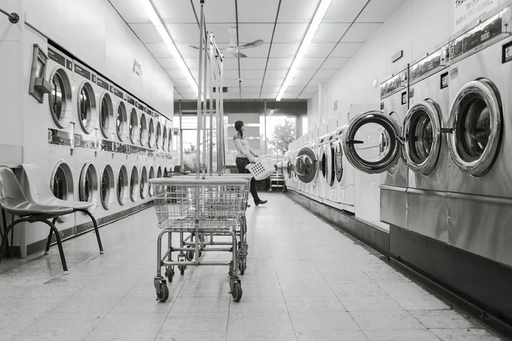 Row of front-loading washing machines in a laundromat with a shopper pushing a cart down the aisle.