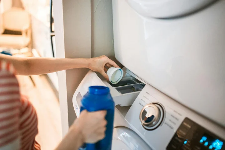 Person loading a front-loading washing machine with a blue detergent bottle.