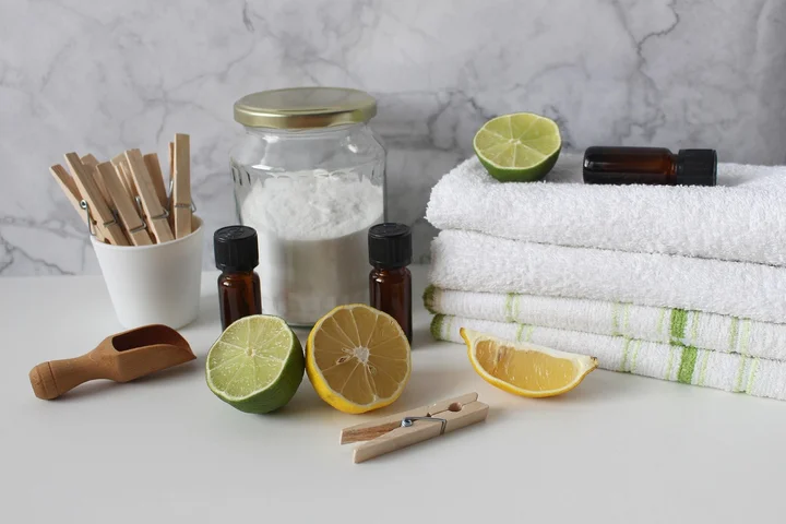 Laundry scene with white towels, citrus halves, wooden clothespins, and cleaning supplies on a marble surface.