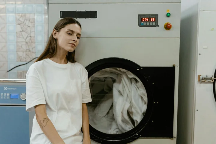 Person wearing a white T-shirt standing beside a front-loading washing machine in a laundromat.