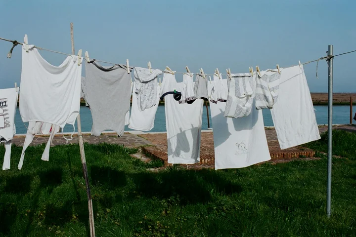 White garments and towels hanging on a clothesline outdoors near a grassy area with a body of water in the background on a sunny day.