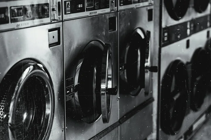 Row of industrial washing machines lined up in a laundromat, representing the evolution of laundry from manual riverbank scrubbing to early soap-based cleaning.
