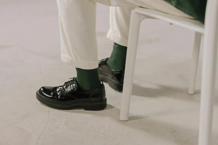 Close-up of polished black leather shoes with green socks and cream pants, resting on a light floor beside a white chair.