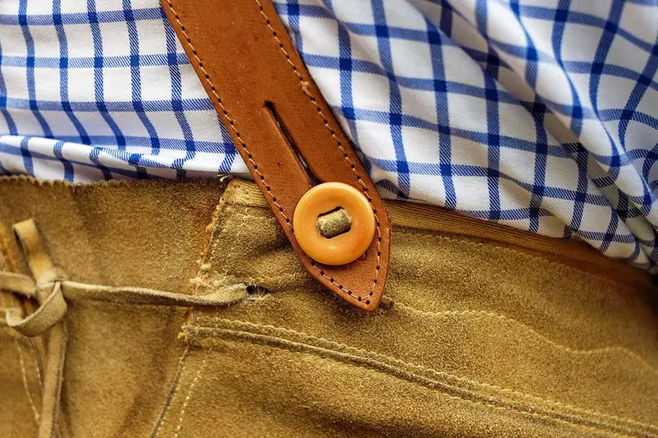 Close-up of a tan leather belt with a circular button and visible stitching resting against a blue-and-white check shirt
