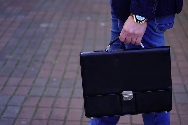 Person in blue jeans and a dark blazer holds a black leather briefcase, standing on a brick-paved sidewalk