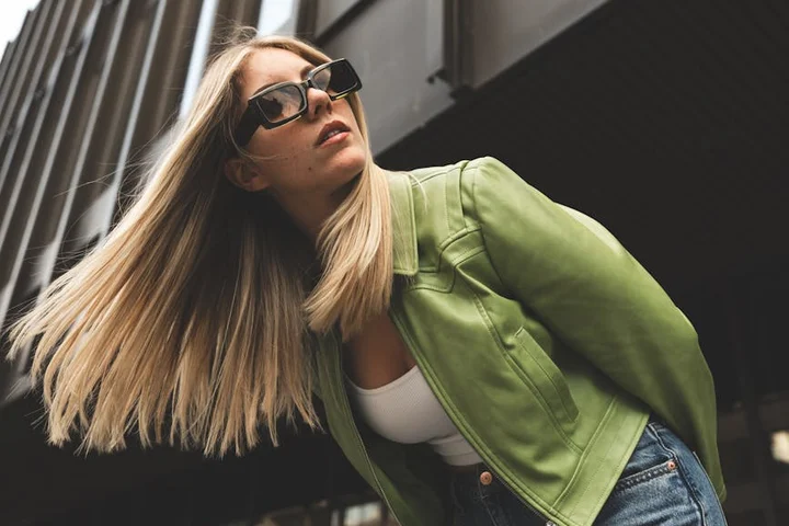 Blonde woman wearing a green jacket and sunglasses, leaning at an angle outdoors, showcasing a casual leather look.
