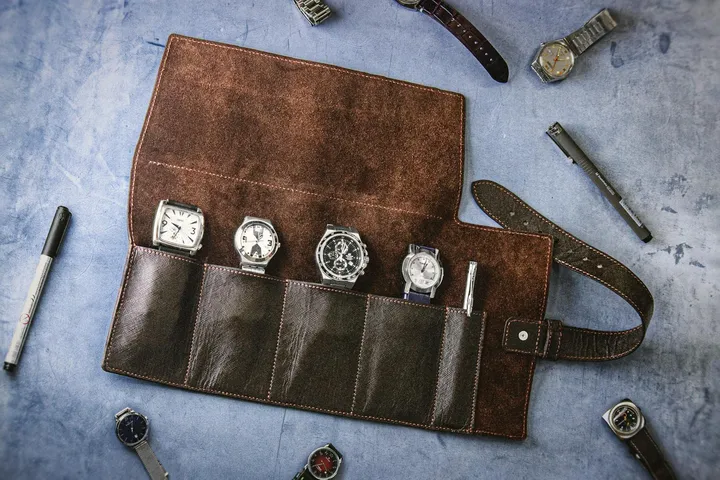 Brown leather watch roll opened to reveal five watches in its pockets, laid on a blue surface with extra straps and tools nearby.