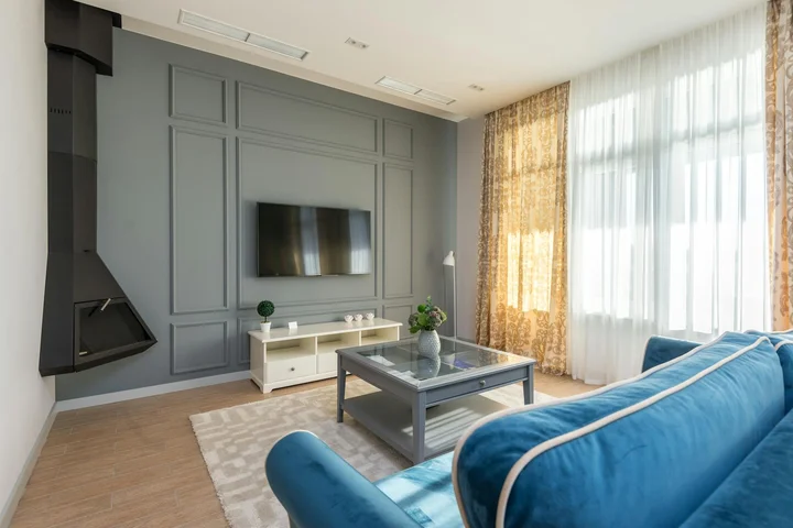 Modern living room with a blue upholstered sofa facing a wall-mounted TV, gray paneled wall, a glass coffee table, and large windows with sheer curtains.