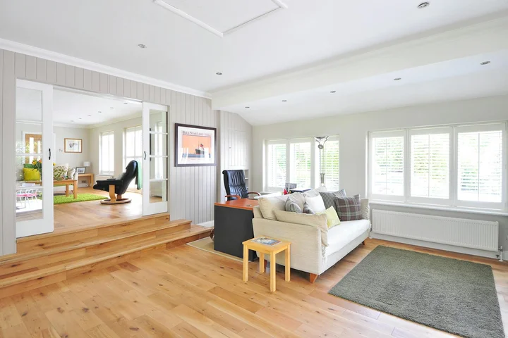 Bright, open living room with light wood laminate flooring, white walls, large windows, a beige sofa, and a small side table.