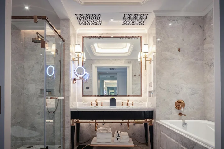 Luxurious bathroom with marble walls and countertops, glass shower enclosure, a large backlit mirror, and a modern dark wood vanity with brass fixtures.