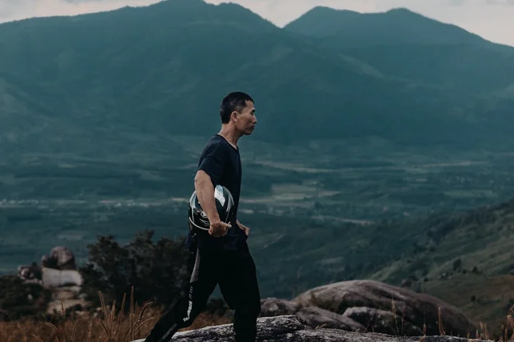 A man stands on a rocky hillside with mountains in the distance.