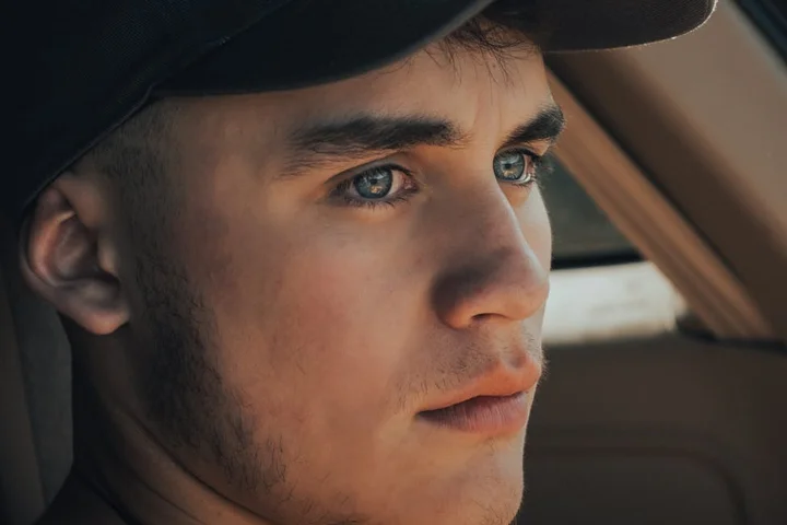 Close-up of a man wearing a baseball cap inside a car, gazing out the window.