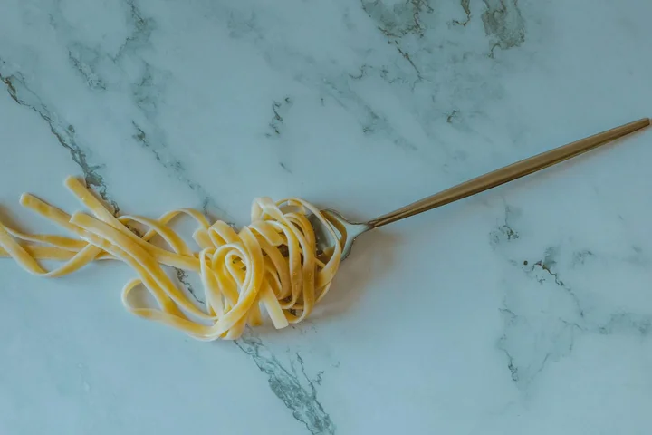 A strand of pasta wrapped around a metal utensil resting on a light blue marble countertop.
