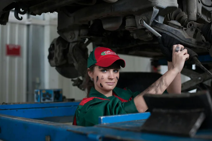 A mechanic in a green uniform and red cap works under a car raised on a lift in a workshop.