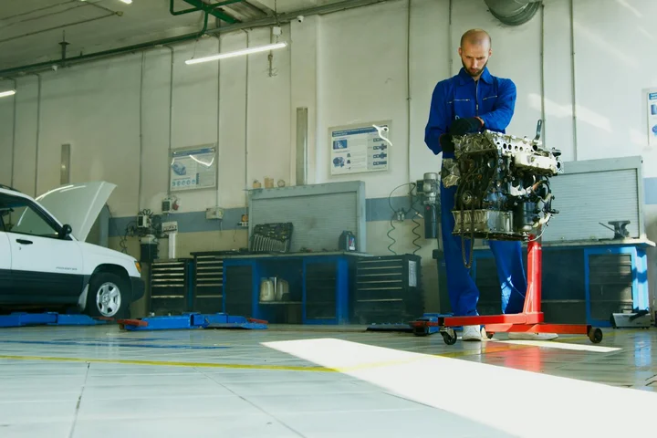 A mechanic in blue coveralls stands beside an engine on a stand in a bright workshop, with tools and a car in the background.