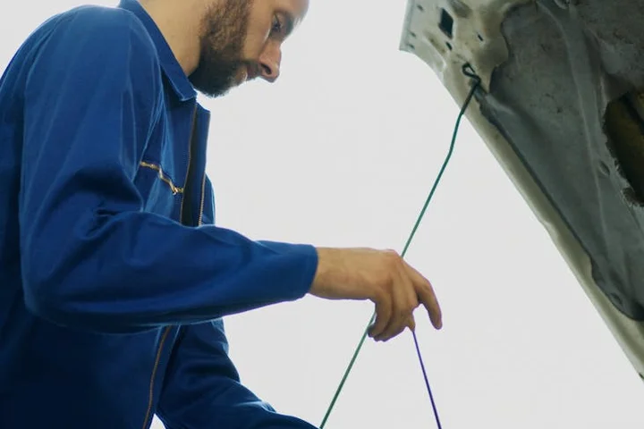 A mechanic wearing blue coveralls leans over a car hood, tightening a component with a tool; the scene suggests grease and oil exposure on durable work fabrics.