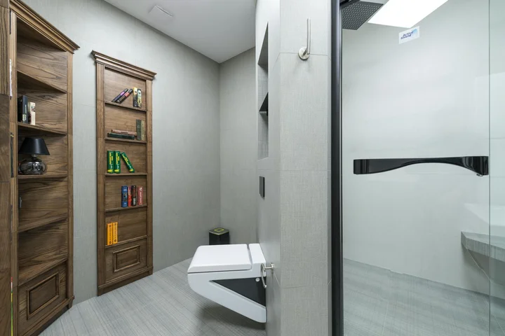 A modern bathroom featuring a wall-mounted toilet, glass shower enclosure, and tall wooden shelves with books.
