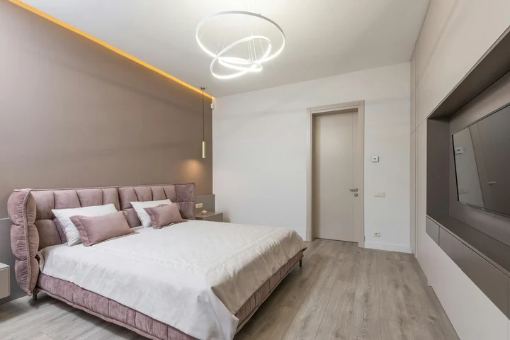 Modern bedroom with a plush mauve headboard bed, white bedding, warm wood flooring, circular ceiling light, and a wall-mounted flat-screen TV.