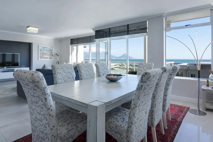 Sunlit modern dining room with a long white table and patterned fabric chairs, large windows, and a view of the outdoors.